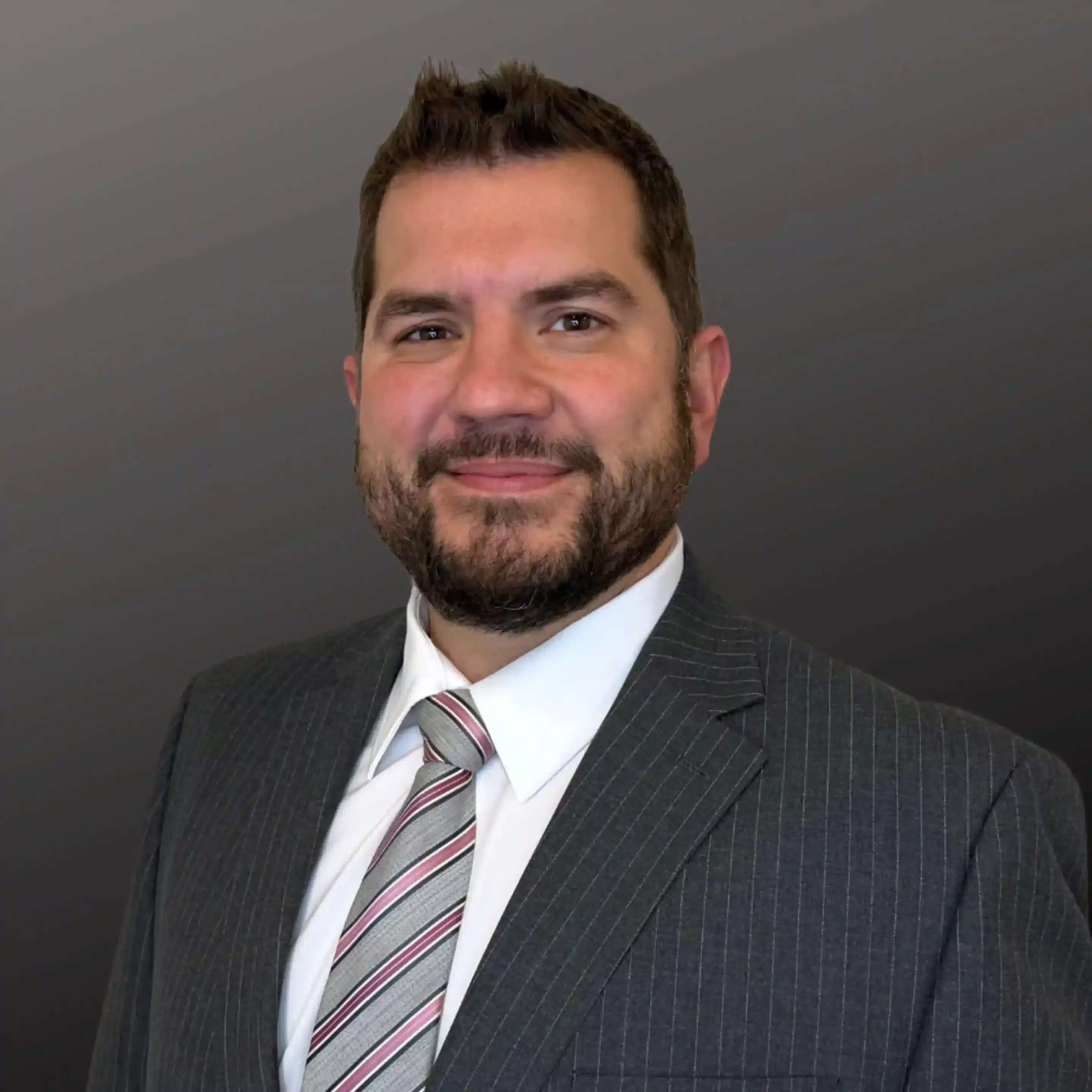 Smiling man in a gray pinstripe suit and striped tie, portrait against a dark backdrop.